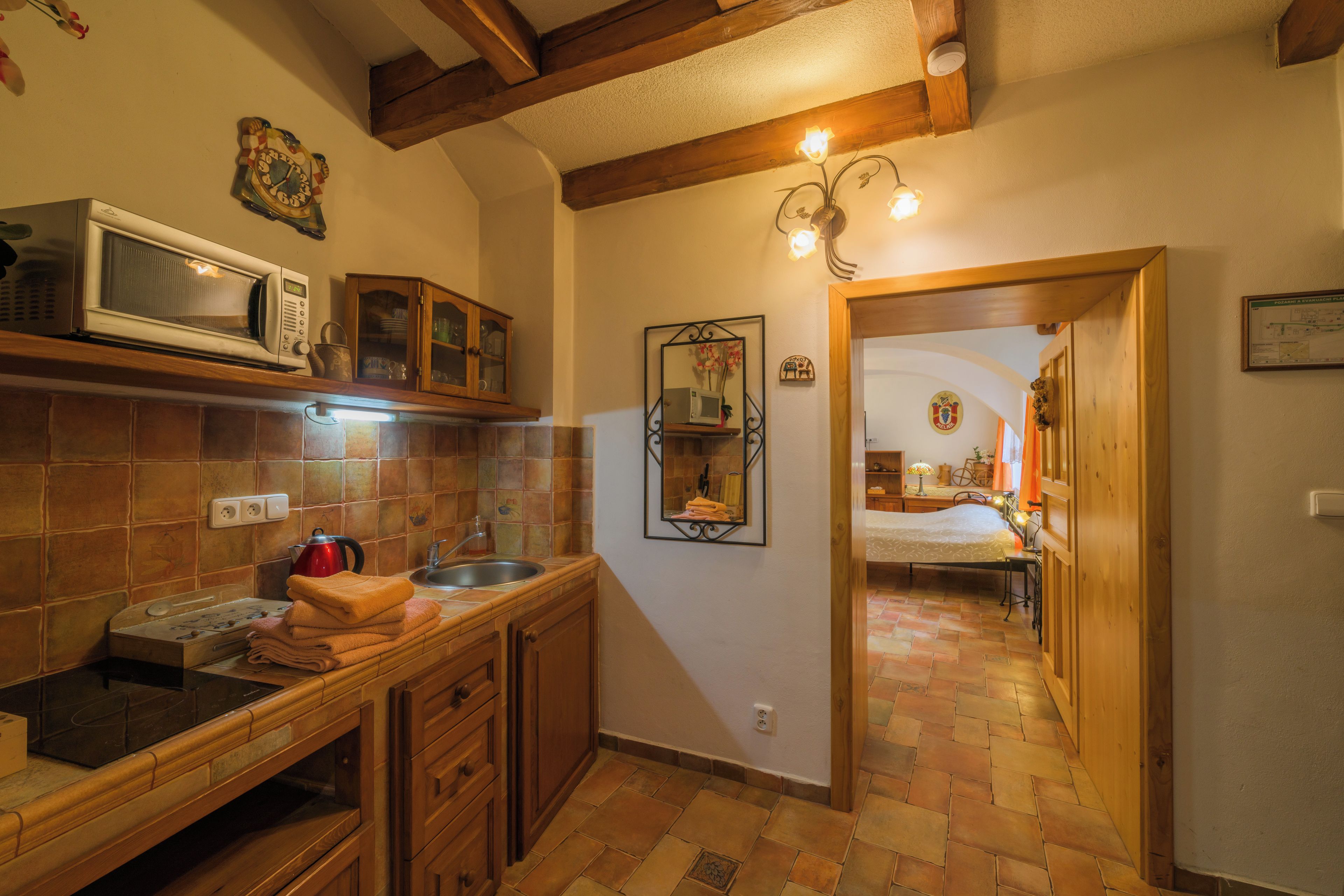 A kitchen with a microwave, cabinets, and a sink. There is a hallway leading to a bedroom with a bed and a dresser.