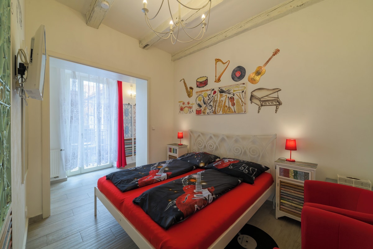 Un dormitorio con cama, una silla roja, una pared con instrumentos musicales y una lámpara roja en la mesita de noche.