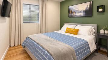 A bedroom with a bed, a painting, a television, and a window with curtains.