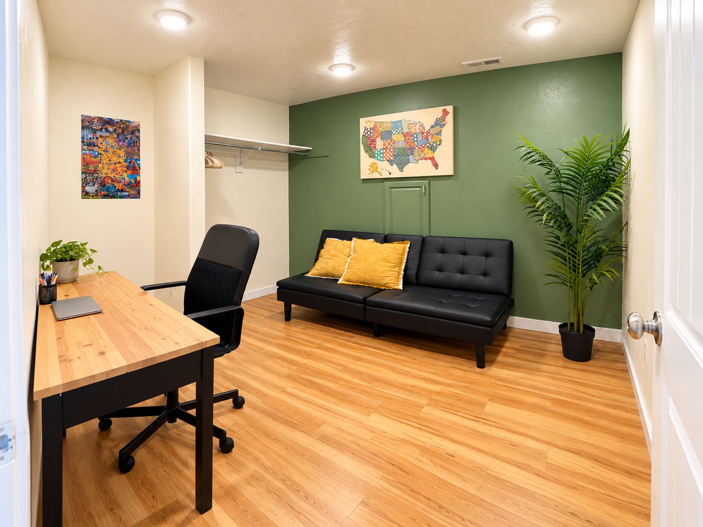 Una habitación con escritorio de madera, silla de oficina negra, sofá negro con cojines amarillos, pared verde con un mapa y una planta en maceta.