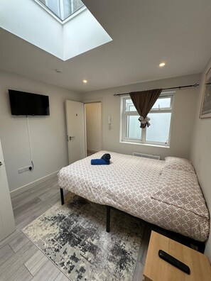 A bedroom with a skylight, a bed with a patterned bedspread, a bedside table, a wall-mounted TV, and a window with curtains. - Studio Sleeps 2 Near Elephant & Castle (London)
