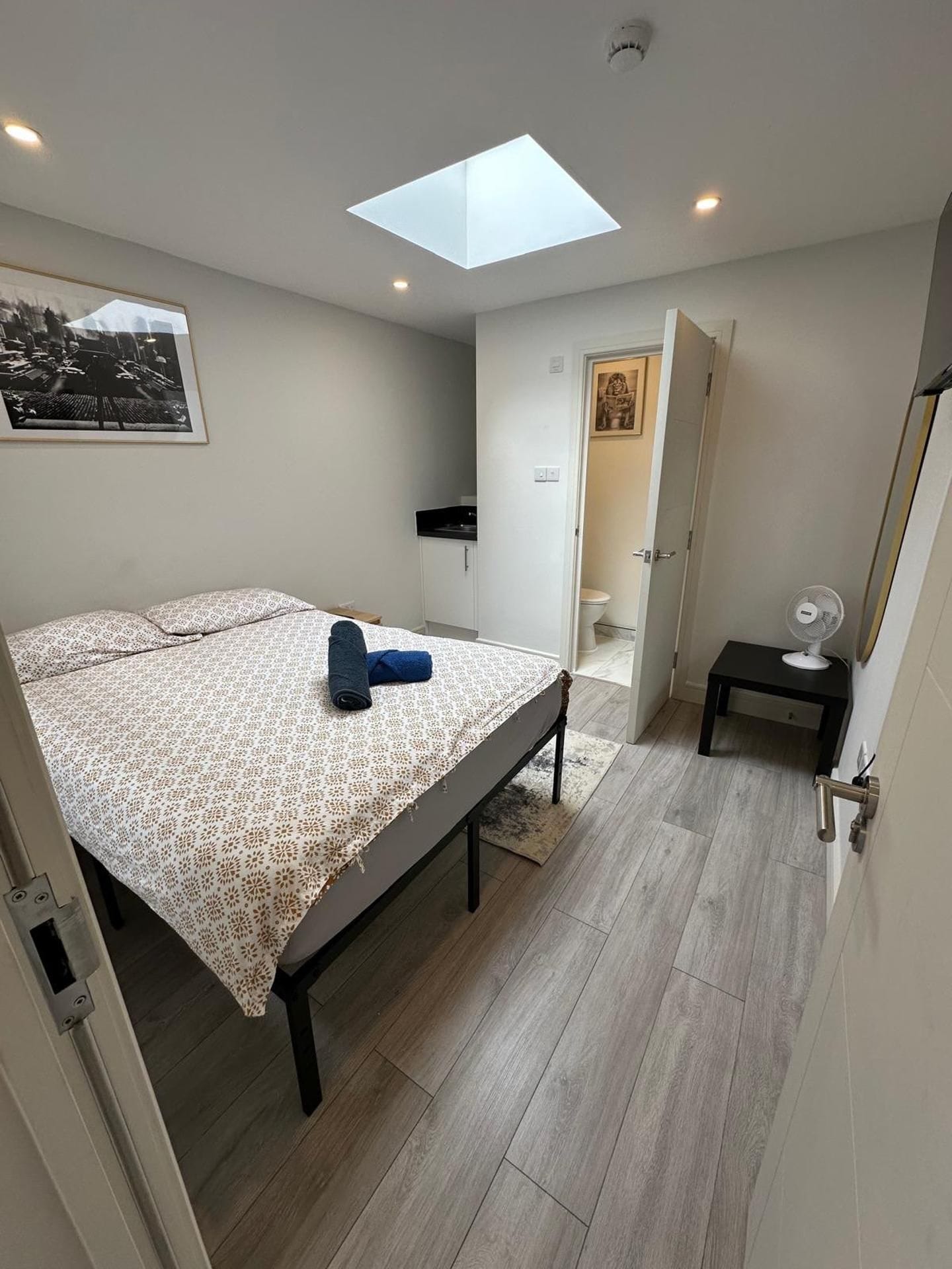 A bedroom with a skylight, a bed with a patterned bedspread, a bedside table with a fan, and a door leading to a bathroom.