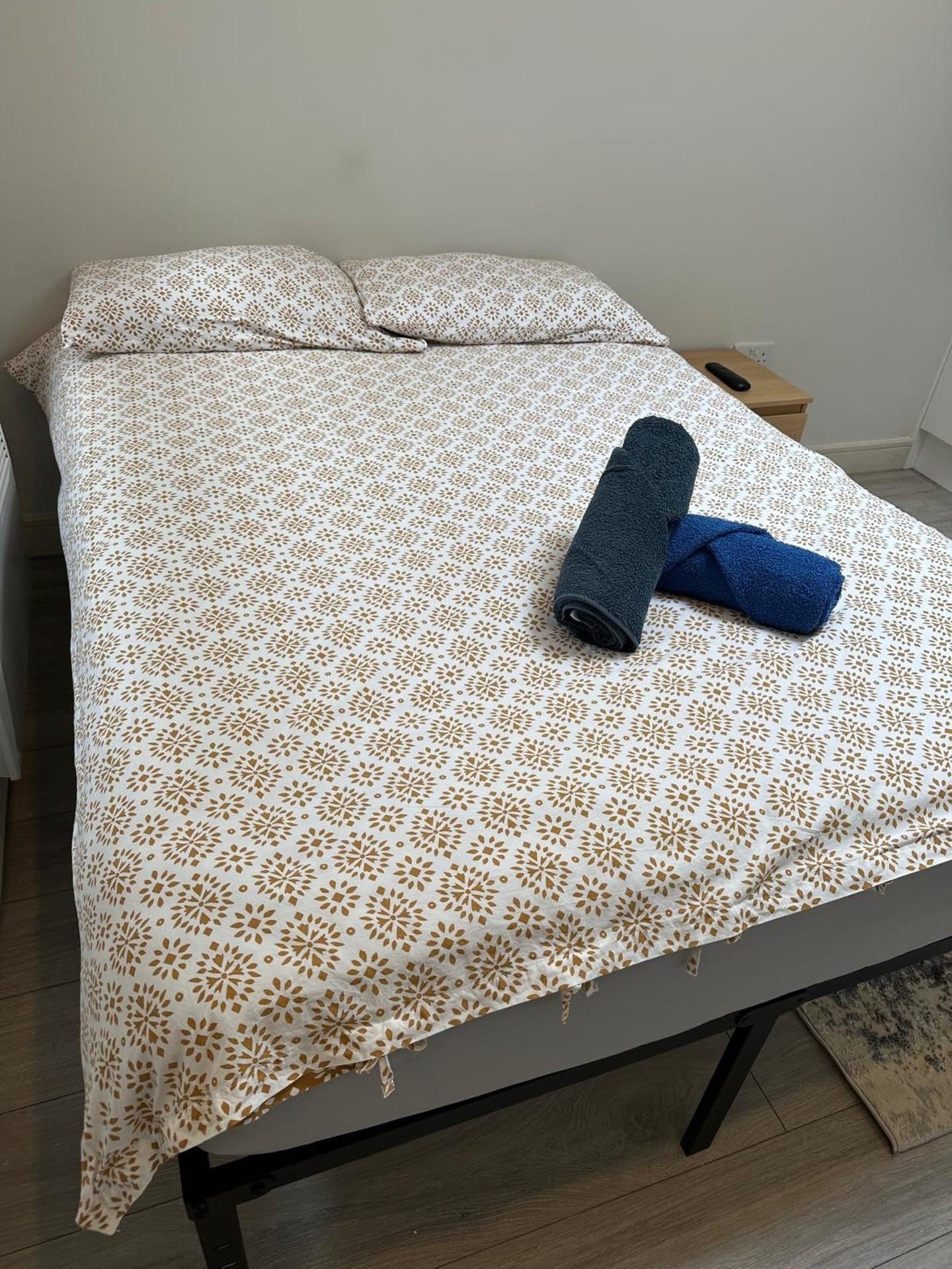 A bed with a patterned bedspread, two rolled towels, and a bedside table with a remote control.