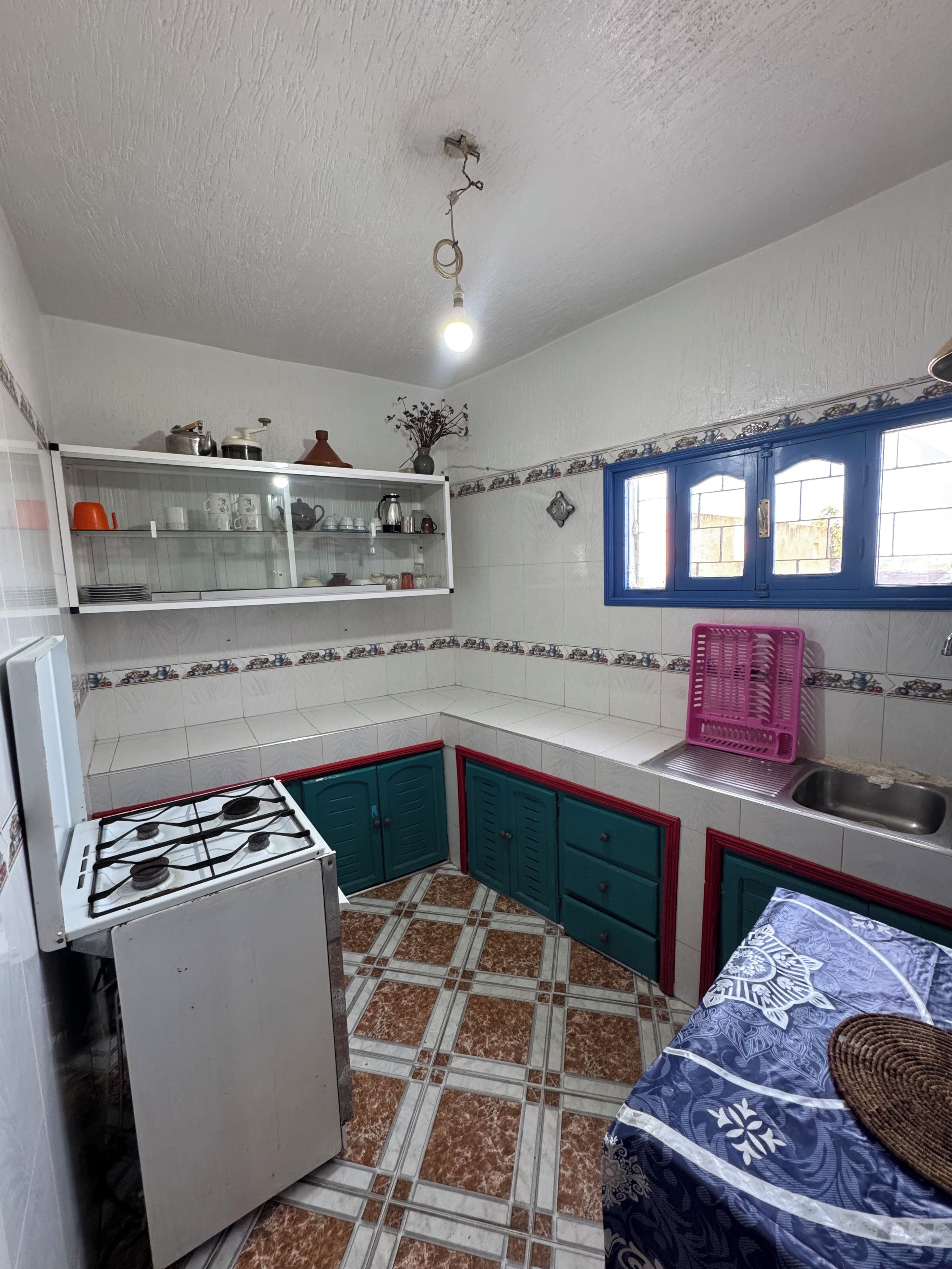Une cuisine équipée d’une cuisinière à gaz, d’un réfrigérateur, d’un évier et d’une fenêtre avec des volets bleus.
