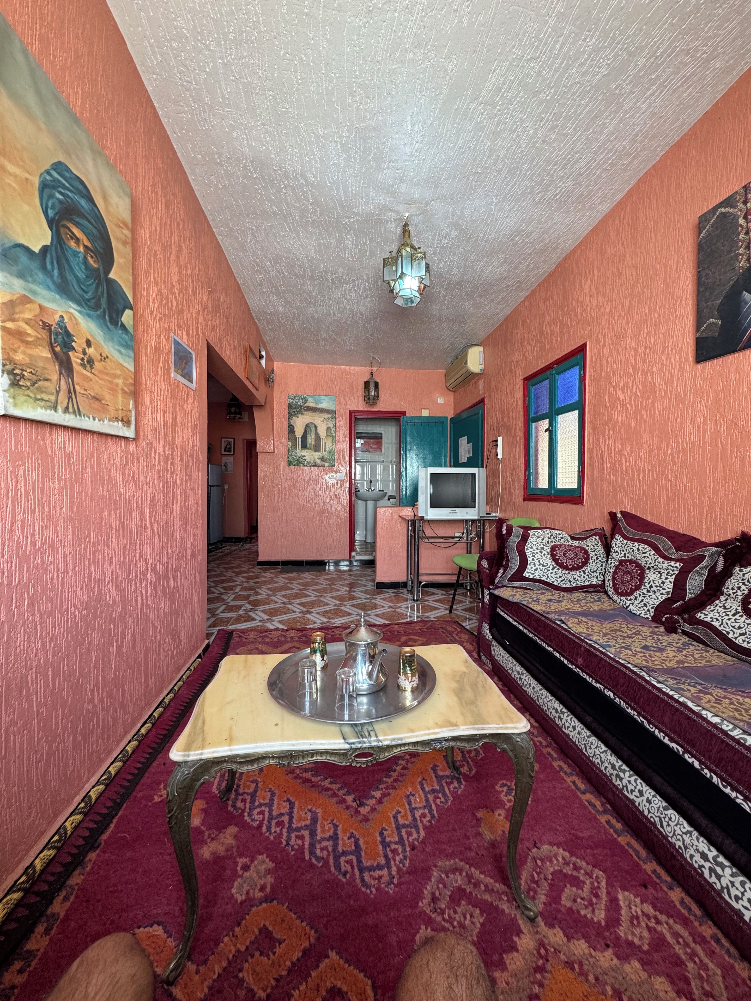 Une pièce avec un tapis à motifs, un canapé agrémenté de coussins décoratifs, une table à manger avec un plateau, et un coin cuisine à l’arrière-plan.