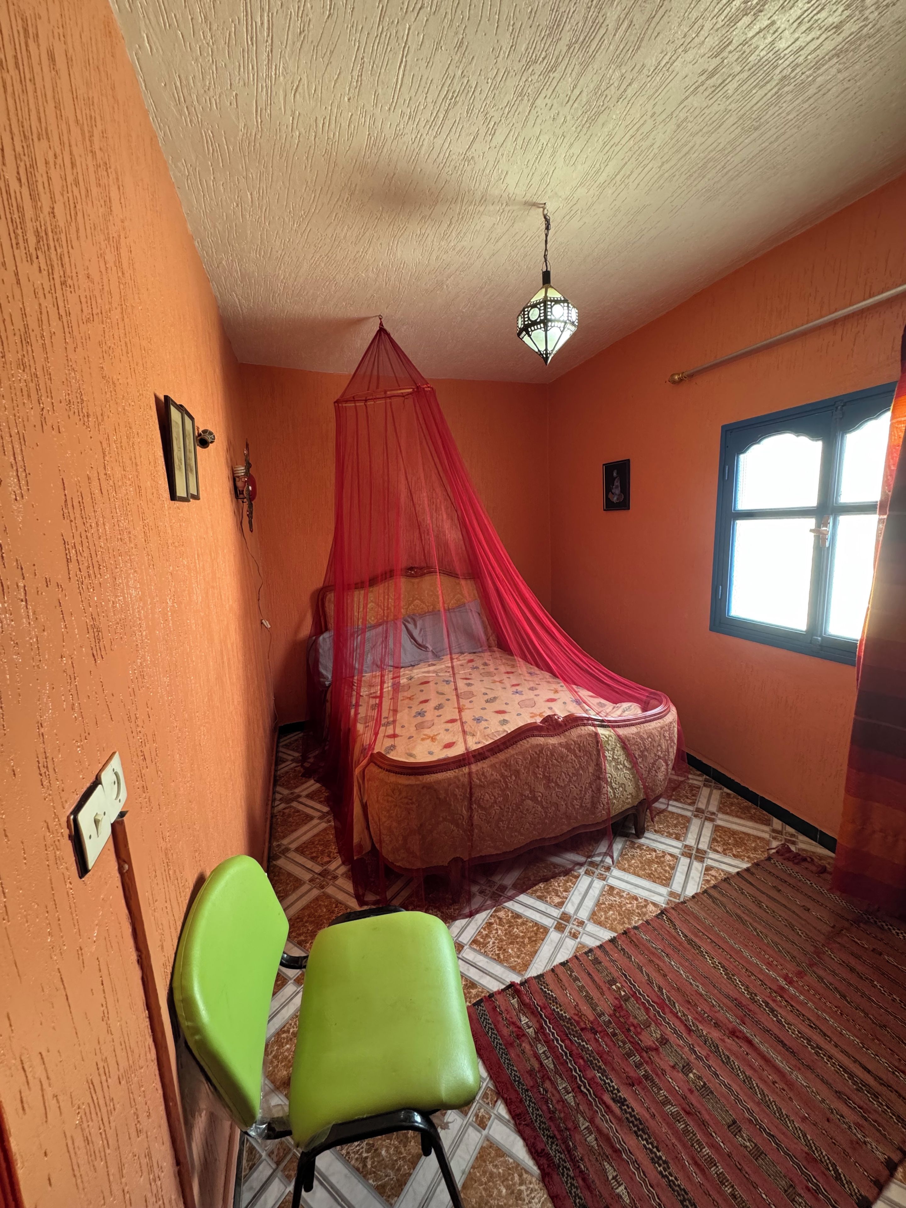 Une chambre avec un lit placé sous un dais rouge, une chaise verte, une fenêtre et un tapis à motifs.