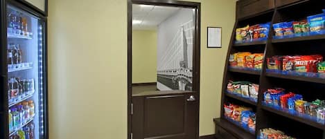 A hallway with a refrigerator stocked with beverages, a door leading to a room with a view of a bridge, and shelves with snacks.