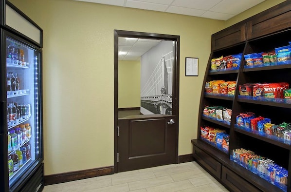 A hallway with a refrigerator stocked with beverages, a door leading to a room with a view of a bridge, and shelves with snacks.