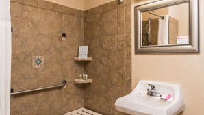 Ein Badezimmer mit Dusche, Waschbecken und Spiegel. In der Dusche befindet sich eine Haltestange, ein Ablagefach mit Pflegeprodukten und ein kleiner Mülleimer.