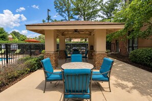 An outdoor patio with a round table and four blue chairs, a covered seating area, and a pool in the background.