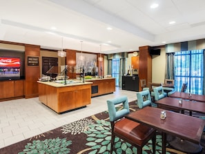 A hotel dining area with a buffet station, seating arrangements, and a television displaying a beverage advertisement.