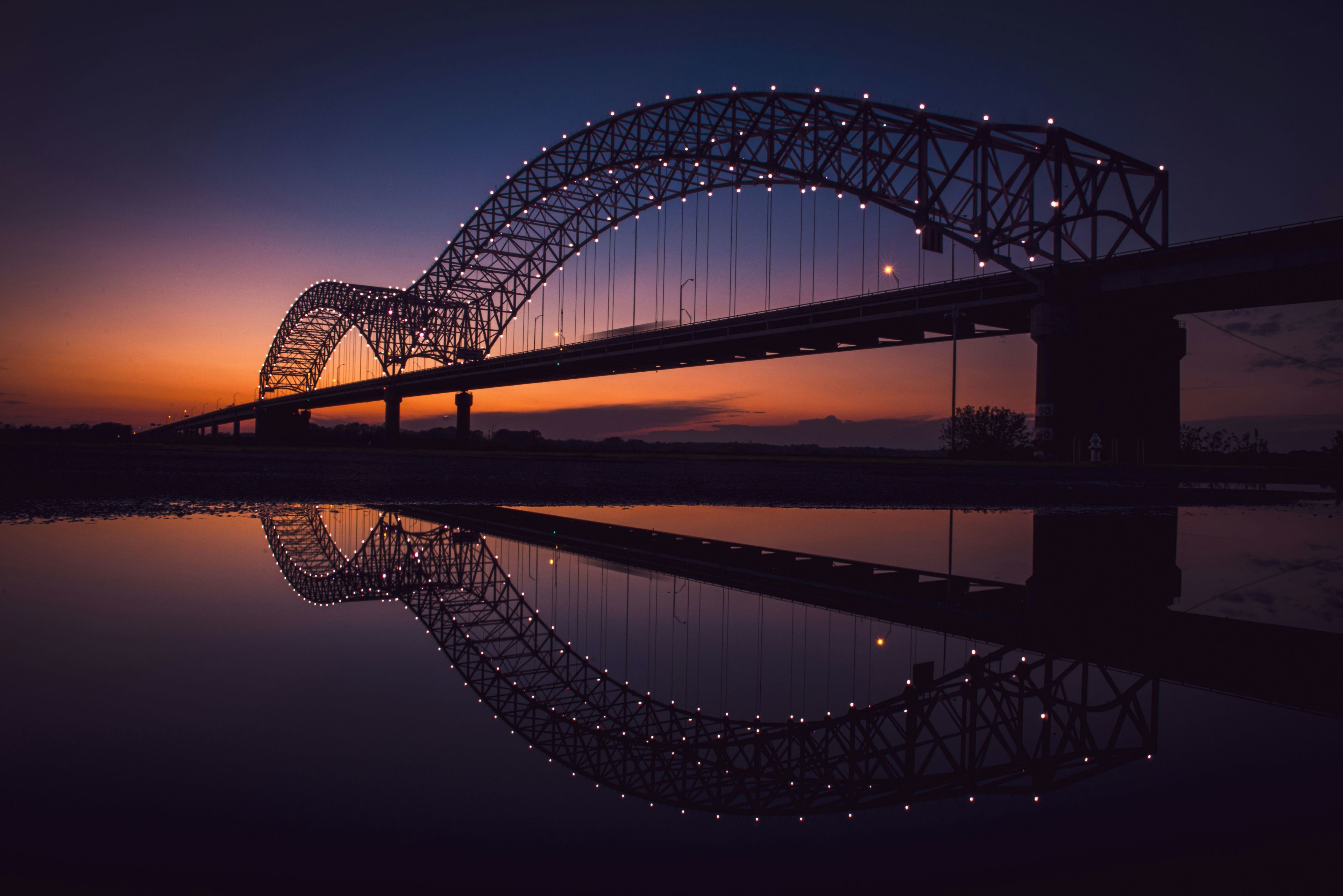 A bridge with a distinctive arch design, spanning over a body of water, with its reflection visible in the water below.