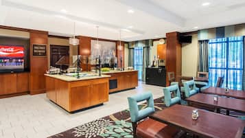 Espaço de refeições de hotel com estação de buffet, mesas e uma televisão exibindo um anúncio de bebida.