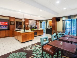 A hotel dining area with a buffet station, seating arrangements, and a television displaying a beverage advertisement.