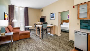Quarto de hotel com sofá, escrivaninha e cadeira, televisão, cama e cozinha equipada com lava-louças.