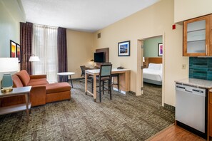 A hotel room with a sofa, a desk with a chair, a television, a bed, and a kitchen area with a dishwasher.