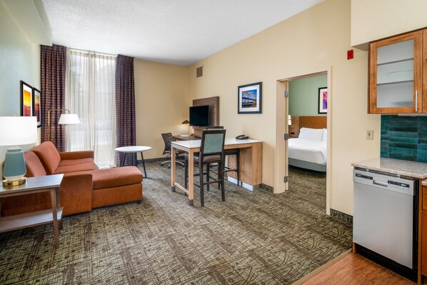 A hotel room with a sofa, a desk with a chair, a television, a bed, and a kitchen area with a dishwasher.