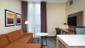 A hotel room with a sofa, a small round table, a desk with a chair, a television, and two framed pictures on the wall.