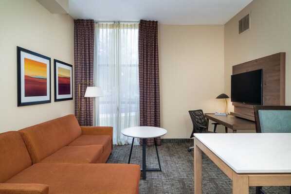 A hotel room with a sofa, a small round table, a desk with a chair, a television, and two framed pictures on the wall.