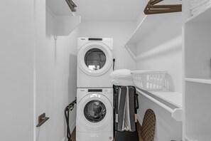 A compact laundry room with two stacked front-loading washers and dryers, a drying rack, and a shelf with a basket. - Beachy Vibes - Gulf Views • Beach Access • Sleeps 12 (Bolivar Peninsula)