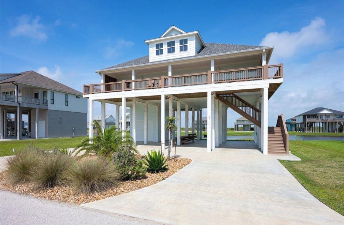 Una casa frente al mar de dos pisos con un porche cubierto, una escalera que conduce a un nivel superior y un jardín delantero con jardines.