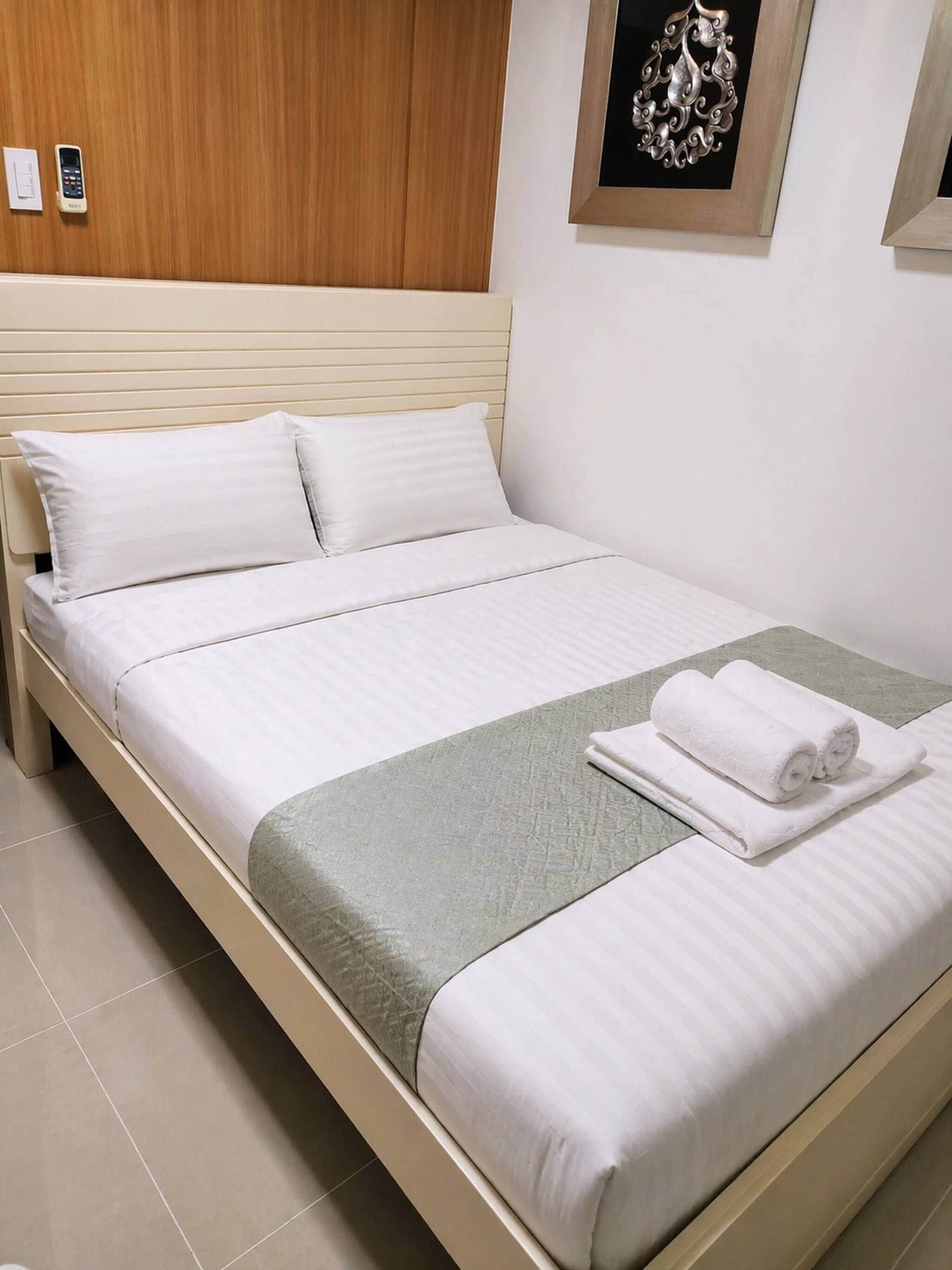 Una cama bien hecha con sábanas blancas, un edredón gris con textura y toallas enrolladas a un lado.