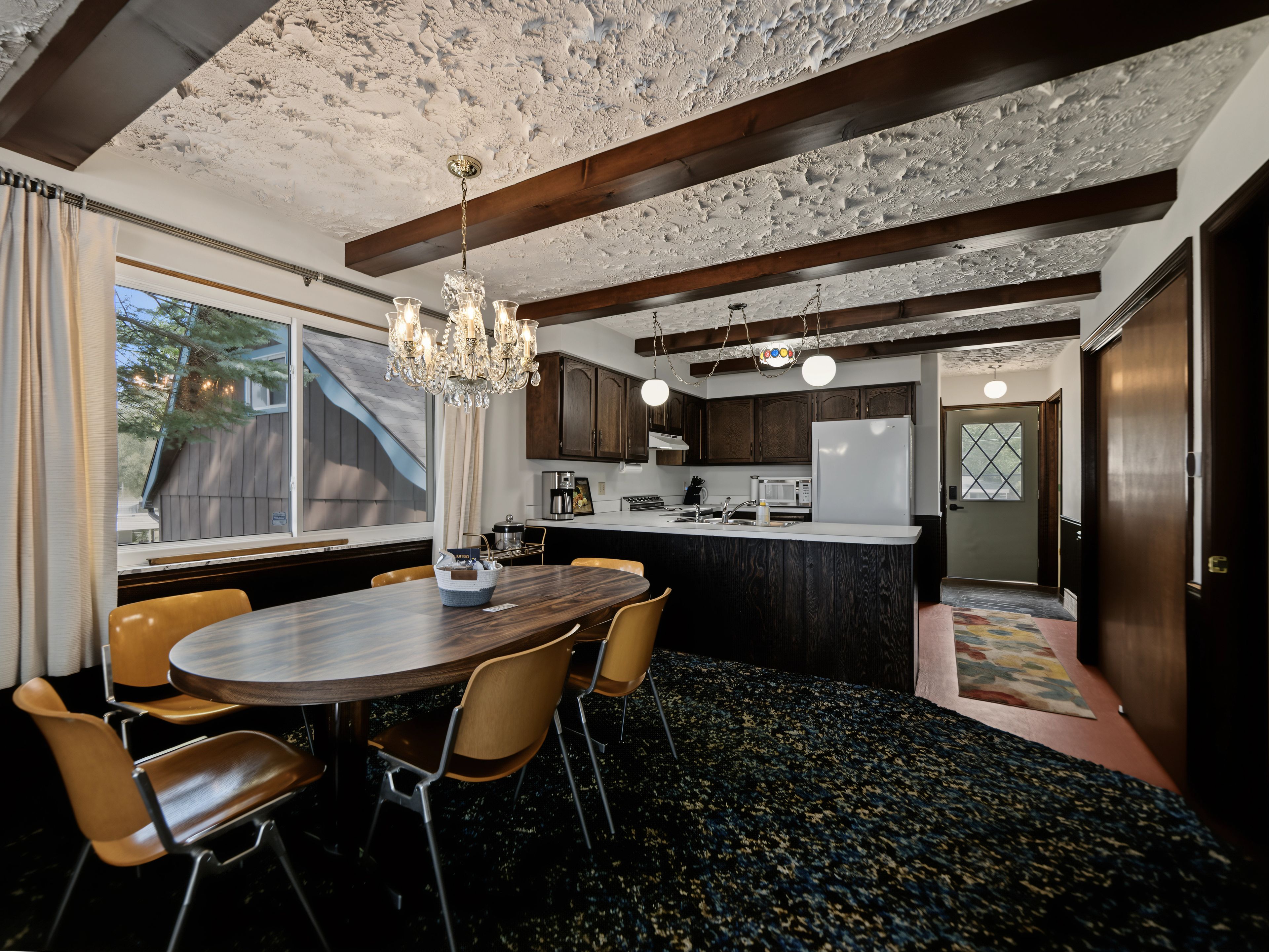 A dining area with a wooden table and chairs, a chandelier, and a kitchen with dark wooden cabinets and a white refrigerator.