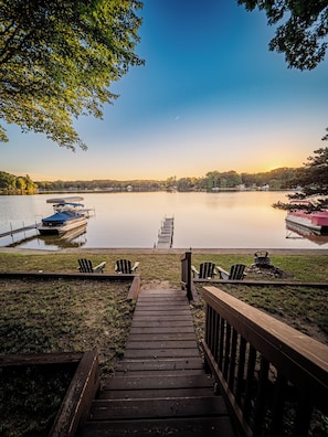 A wooden dock leading to a calm lake with boats docked on the left, a grassy area with Adirondack chairs, and a clear sky.