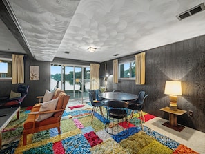 A room with a large window, a round table with chairs, a colorful rug, and a lamp.