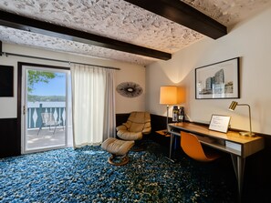 A hotel room with a view of the lake, a desk with a lamp, a chair, and a framed picture on the wall.