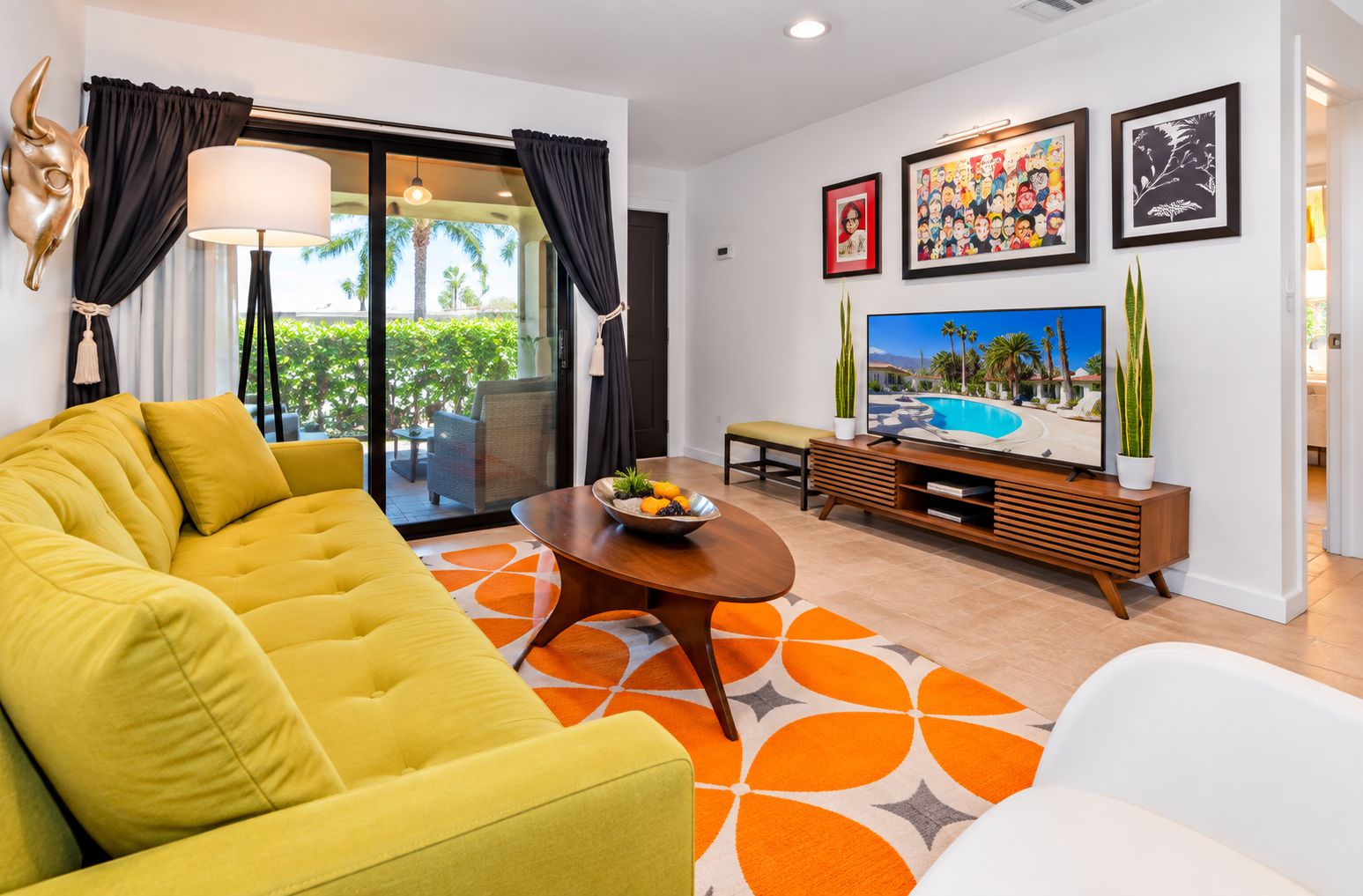 Una sala de estar moderna con un sofá amarillo, una mesa de centro con fruta, un televisor y vista a una piscina.