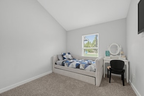 A bedroom with a bed, a desk, a chair, a window, and a wall-mounted mirror.