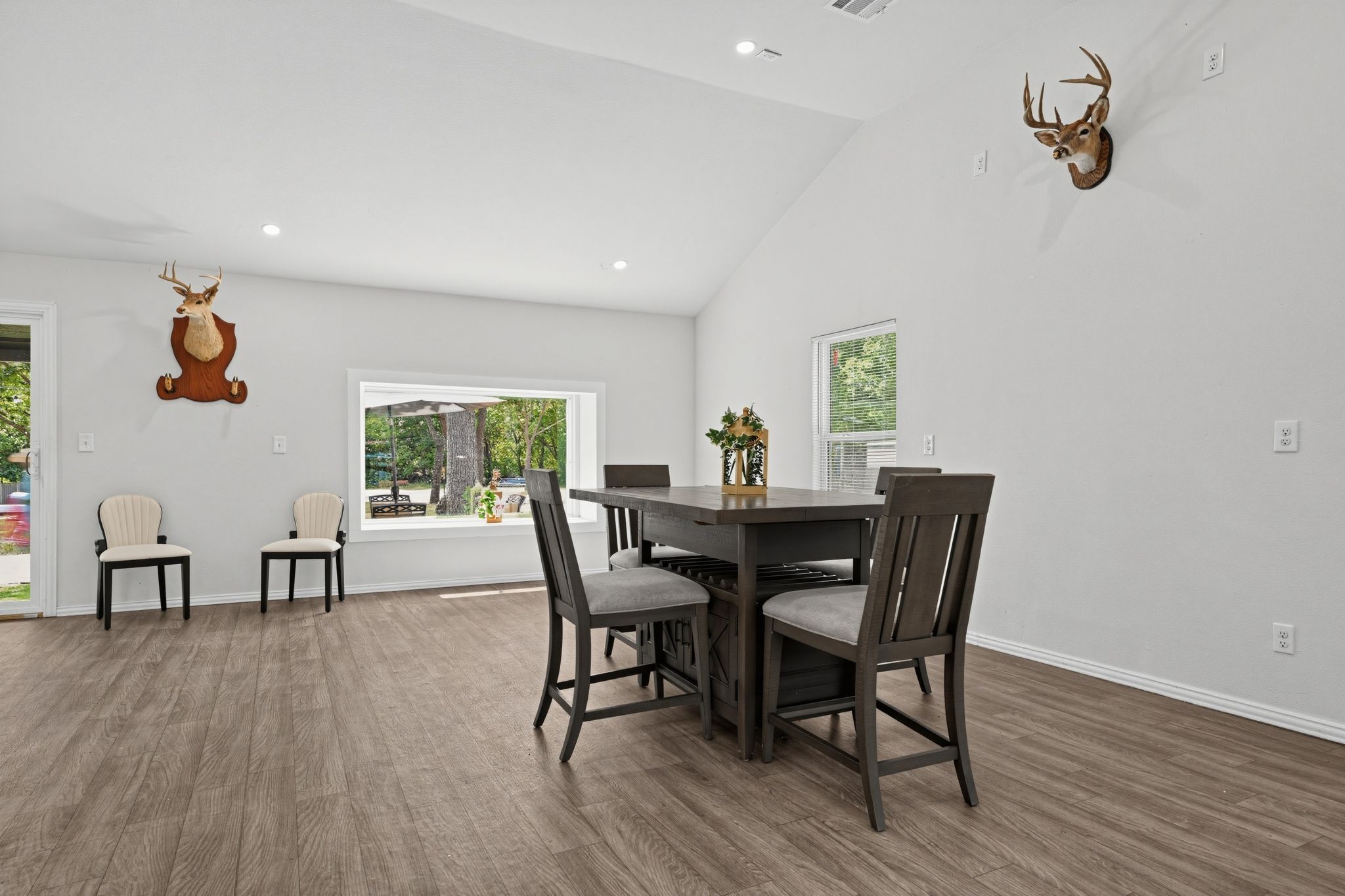A modern dining area with a wooden table and chairs, a mounted deer head on the wall, and a window with a view of greenery.