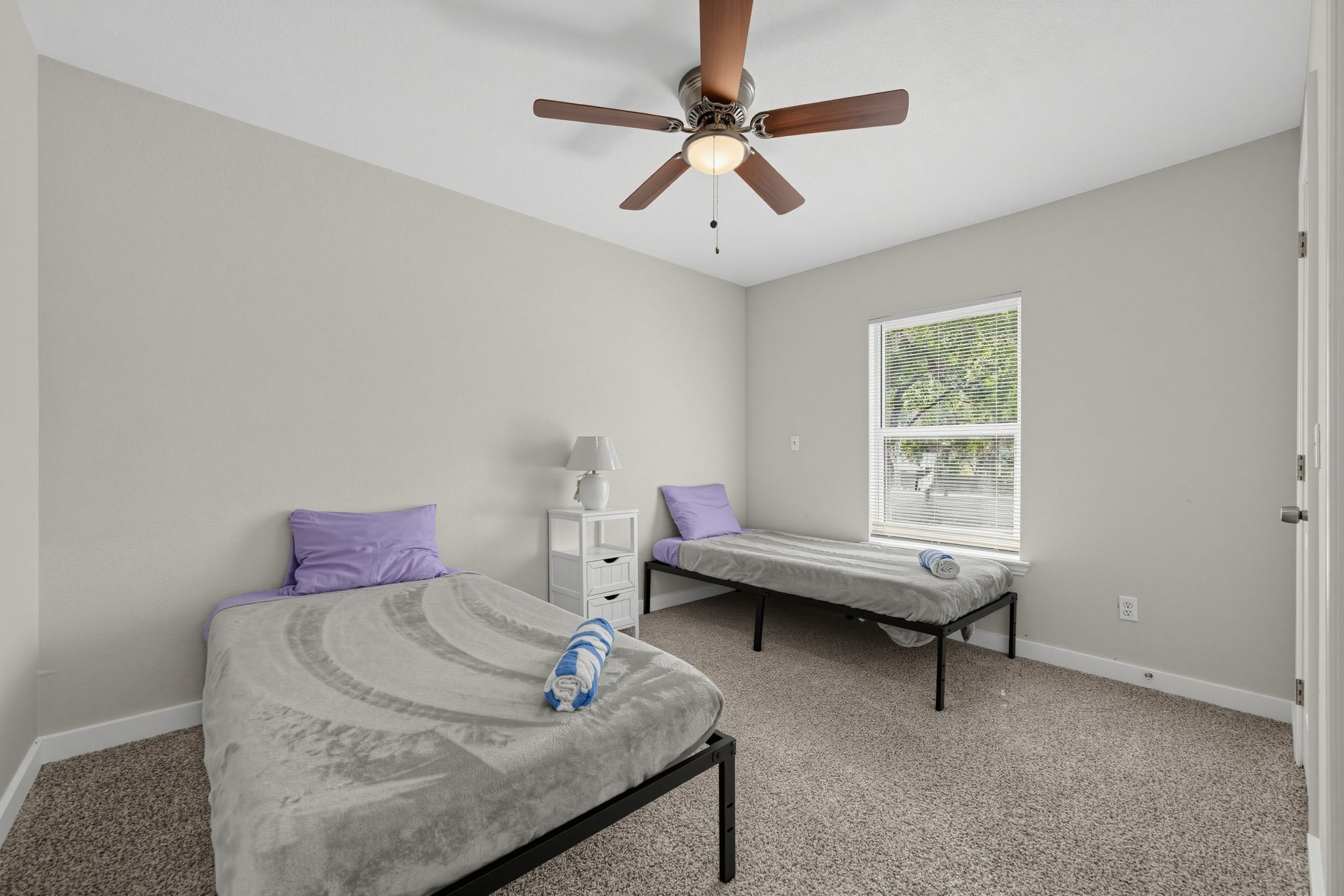 Une chambre avec deux lits simples, un ventilateur de plafond, une table de chevet et une fenêtre avec des stores.