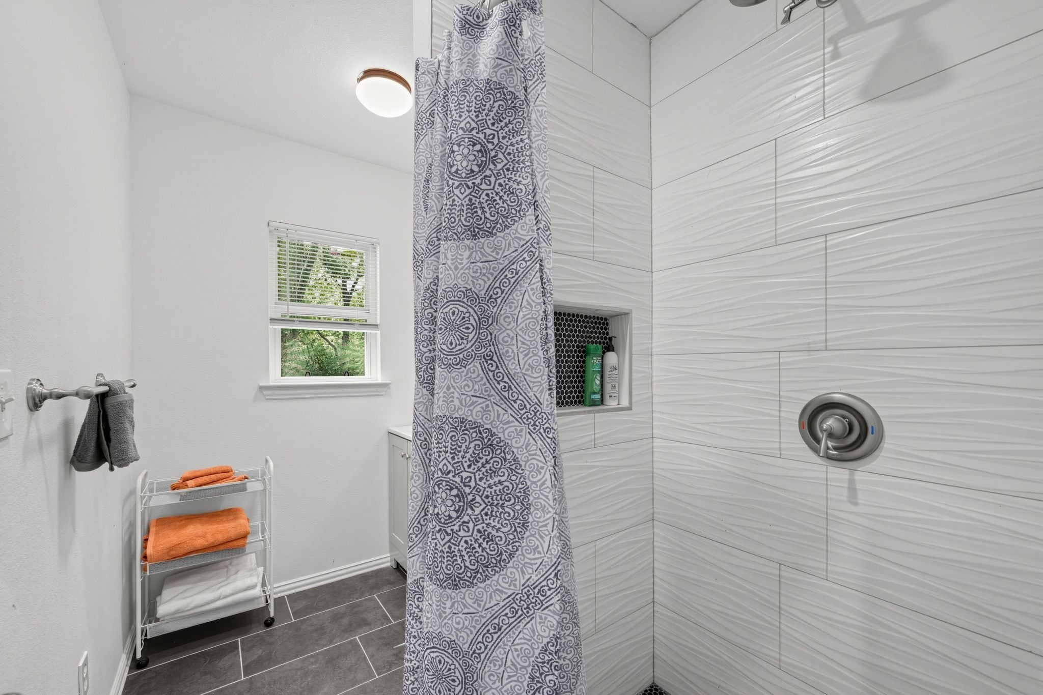 A bathroom with a shower curtain, a towel rack with towels, a window, and a shelf with toiletries.