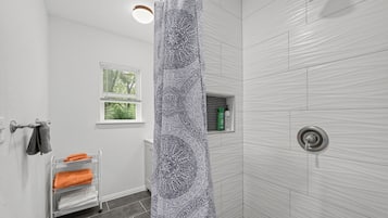 A bathroom with a shower curtain, a towel rack with towels, a window, and a shelf with toiletries.