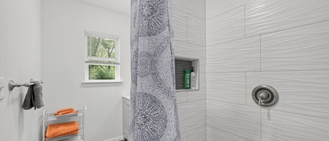 A bathroom with a shower curtain, a towel rack with towels, a window, and a shelf with toiletries.