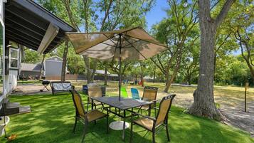 Une terrasse extérieure avec une table et des chaises, entourée d’arbres et d’une pelouse verdoyante.