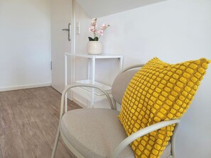 A room with a white chair, a small white table, and a yellow textured pillow. - Heiligenhafenbude 2 (Heiligenhafen)