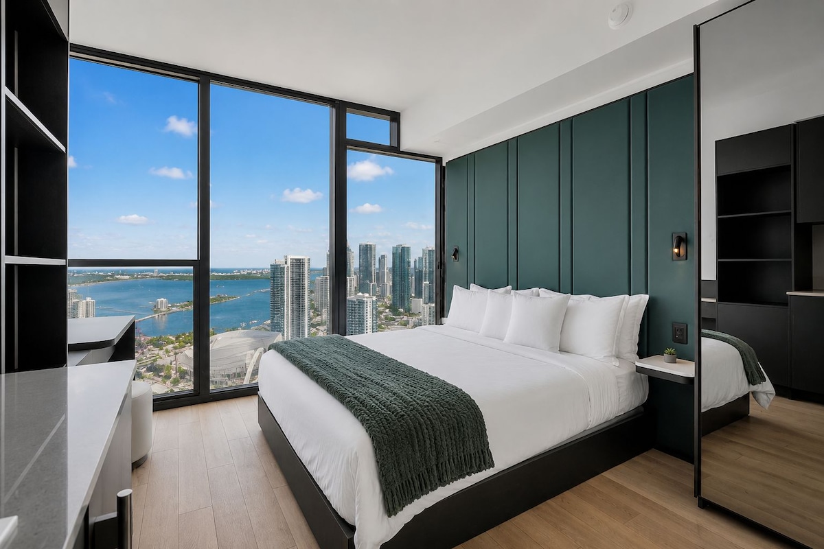 A modern hotel room with a large bed, a desk, and a view of a cityscape through floor-to-ceiling windows.