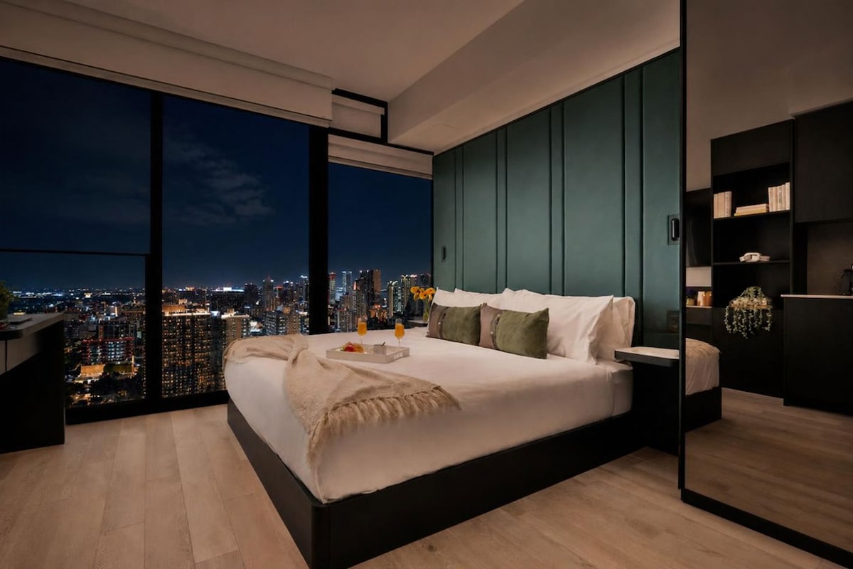 Quarto moderno com vista para a cidade, uma cama grande, mesas de cabeceira e uma estante embutida.