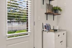 A room with a white door, a window with horizontal blinds, a small cabinet with drawers, and a shelf with a plant and books.