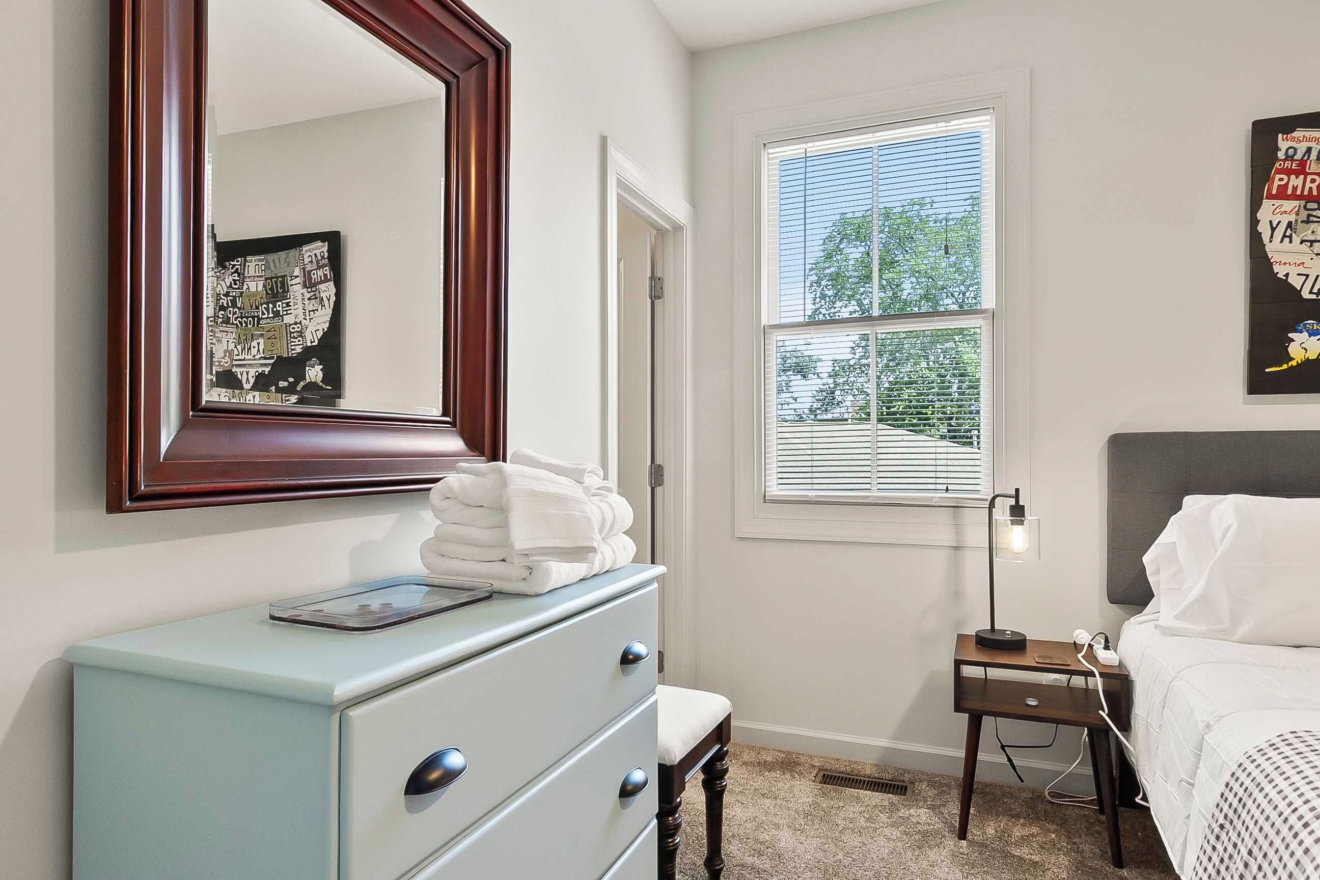 A bedroom with a dresser, a bedside table with a lamp, a bed, a window with blinds, and a wall-mounted mirror.
