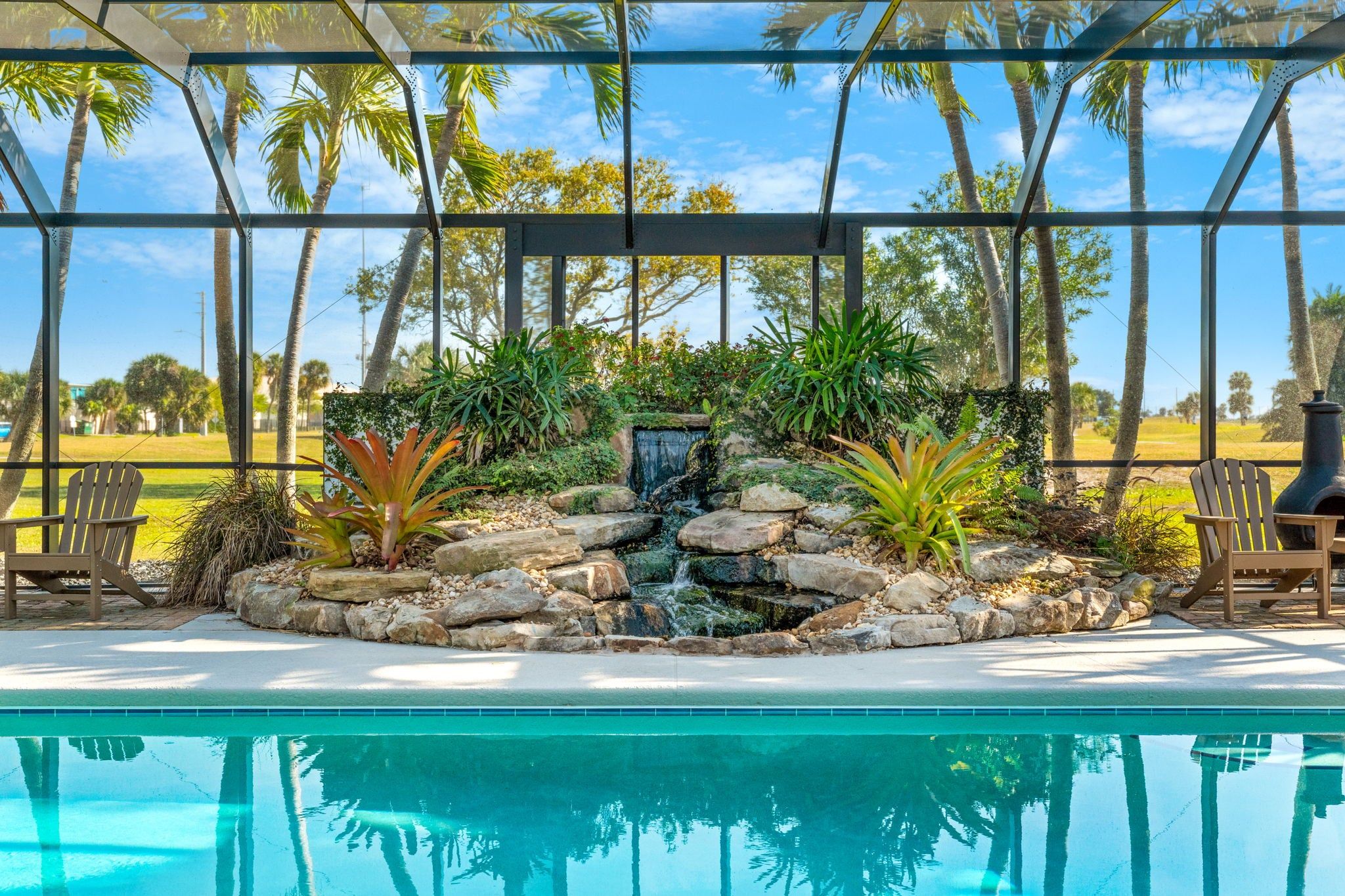 Zona de piscina con cascada, rodeada de plantas tropicales y palmeras.