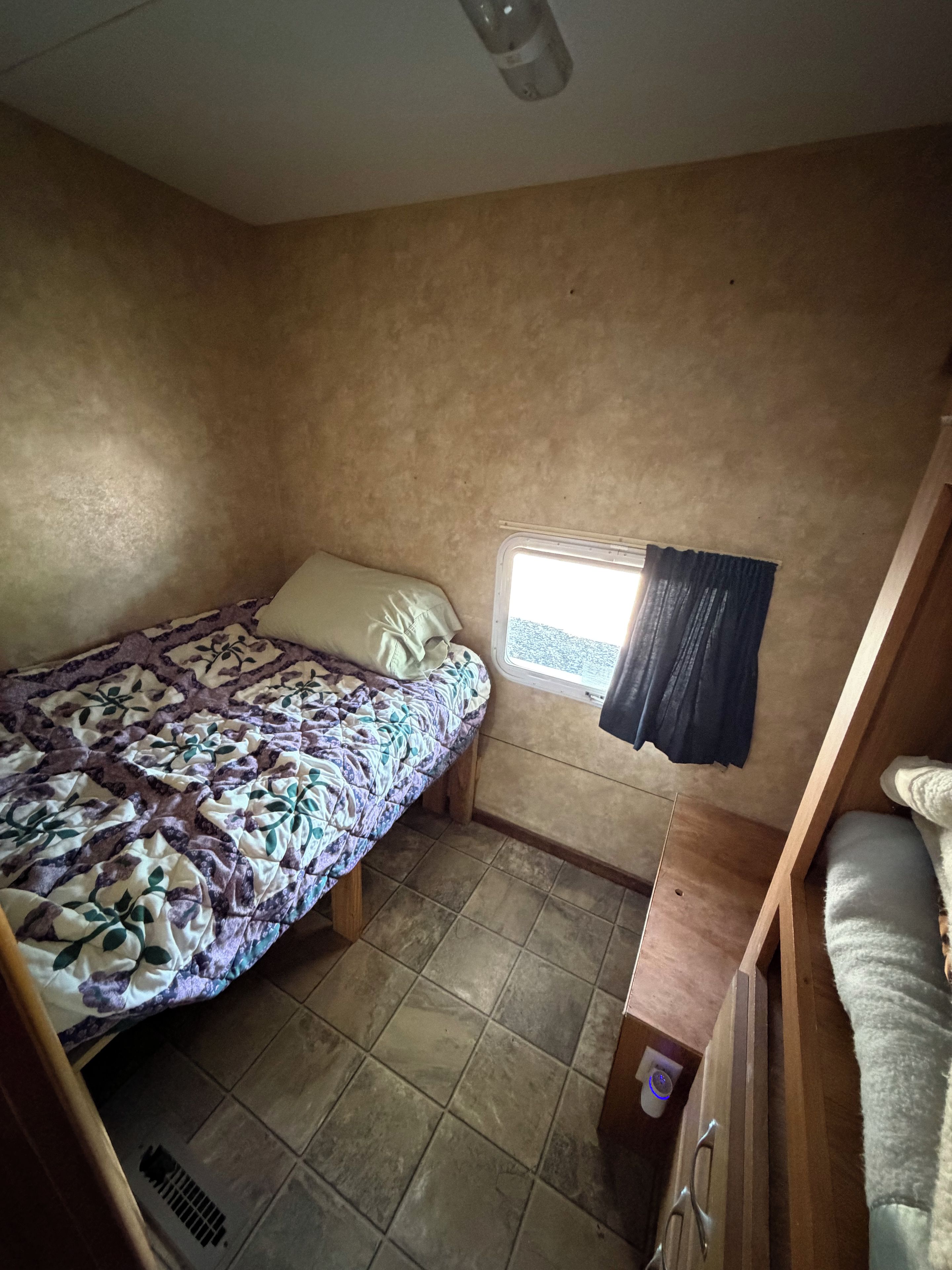 A compact room with a bed, a small window with curtains, a radiator, and a wooden bench.