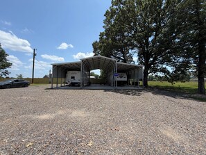 A covered parking area with a gravel surface, a parked car, and a large tree to the right. - Cozy 1-bedroom recreational vehicle in perfect Delight with AC (Delight)
