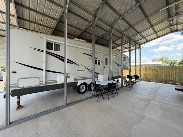 A covered RV parking area with a white RV, outdoor seating, and a wooden fence in the background.