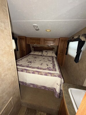A compact RV interior with a bed, a small kitchenette, and a bathroom.