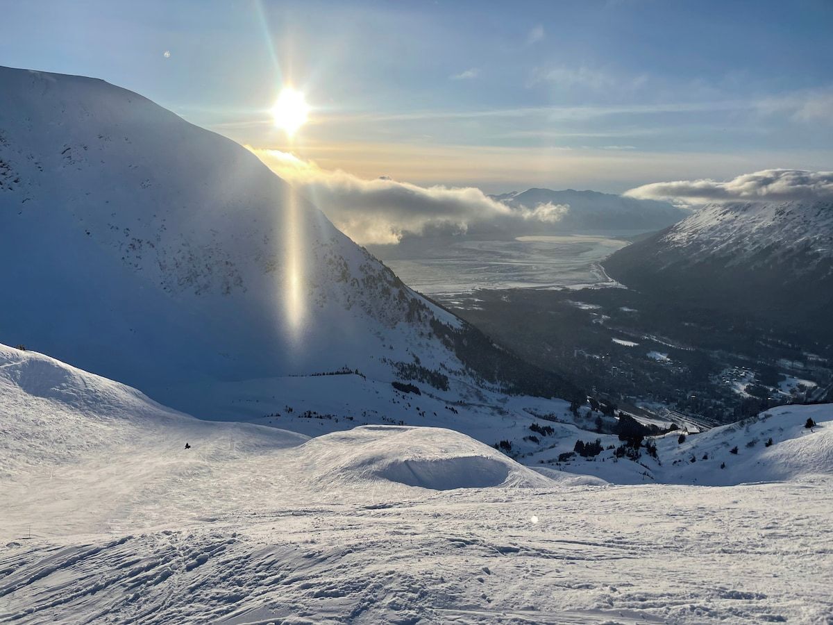 A snow-covered mountain landscape with a clear sky and the sun shining from behind the peaks.