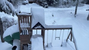 Eine mit Schnee bedeckte Holzspielkonstruktion mit Rutsche und Schaukeln, umgeben von schneebedeckten Bäumen und einem Fahrzeug im Hintergrund.